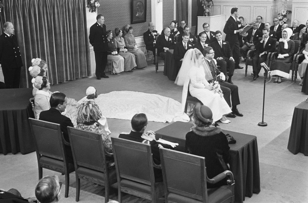 Schwarz-weiß-Foto einer Hochzeitszeremonie mit sitzenden und stehenden Gästen, einem Mann mit Mikrofon, einer Frau mit Blumenstrauß, einem Podium mit Mikrofon, Blumenschmuck, einem Vorhang, einem gerahmten Foto an der Wand und einer Tür im Hintergrund.