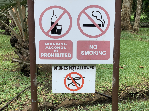 Schild am Straßenrand mit der Aufschrift "Alkoholgenuss ist untersagt" und "Rauchen verboten" mit Bäumen und Gras im Hintergrund.