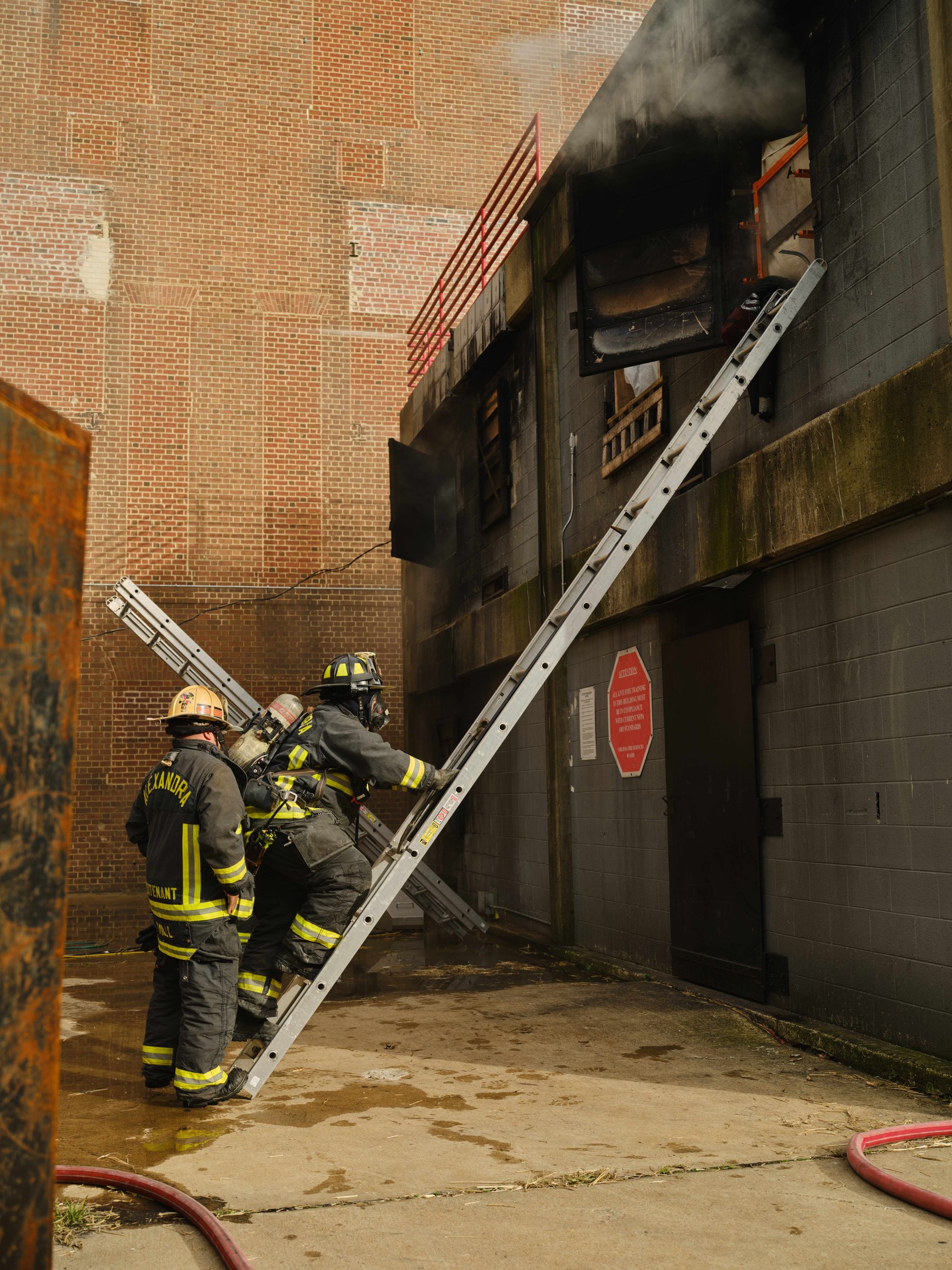 Feuerwehrleute mit Helmen klettern an einer Leiter zu einem raucherfüllten Gebäude hoch, mit einem Metallobjekt links und Rohren am Boden.