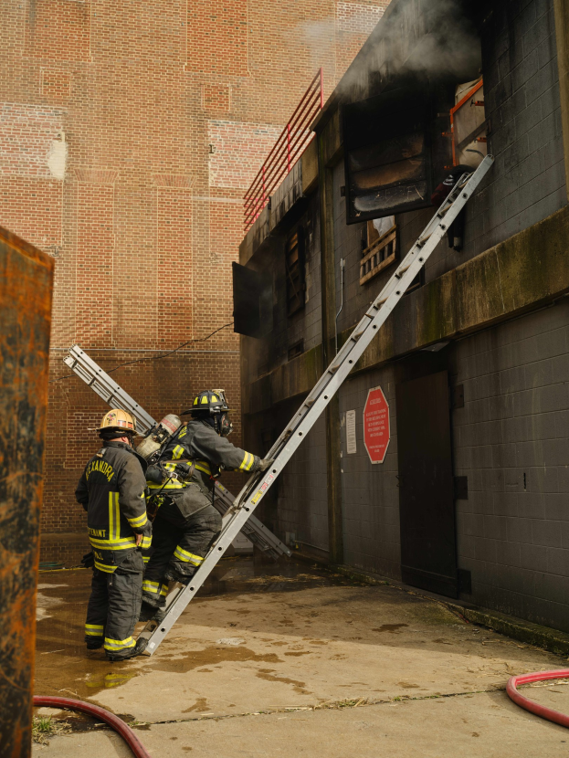 Feuerwehrleute mit Helmen klettern an einer Leiter zu einem raucherfüllten Gebäude hoch, mit einem Metallobjekt links und Rohren am Boden.