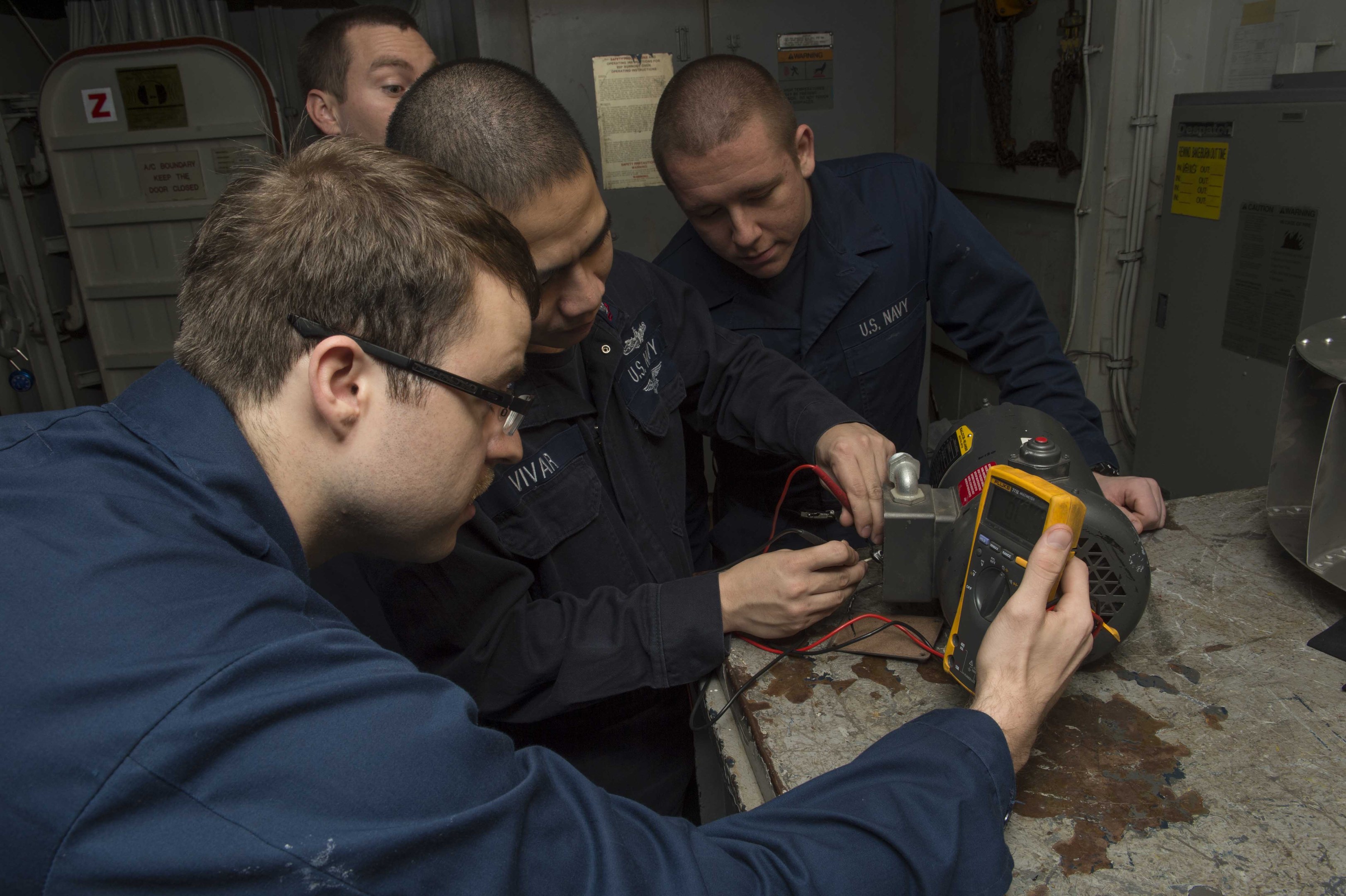 Vier Männer in einem Raum, die an einer Maschine arbeiten, mit einem Tisch mit Gegenständen rechts und Maschinen und Papieren an den Wänden im Hintergrund.