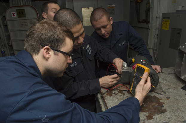 Vier Männer in einem Raum, die an einer Maschine arbeiten, mit einem Tisch mit Gegenständen rechts und Maschinen und Papieren an den Wänden im Hintergrund.