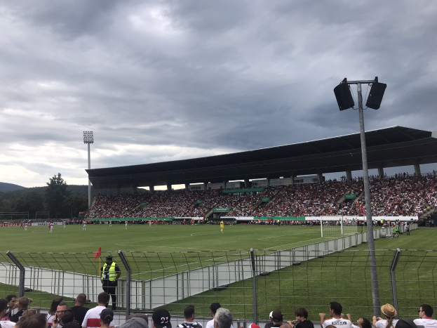 Große Zuschauermenge bei einem Fußballspiel in einem bewölkten, offenen Stadion mit beleuchtetem Feld, eingezäuntem Gelände und fernen Hügeln.