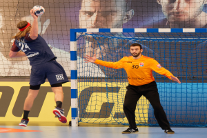 Zwei Männer beim Handballspielen vor einem Tor mit Netz und einem Banner mit Text und Bildern von Menschen im Hintergrund, beide tragen sportliche Schuhe.