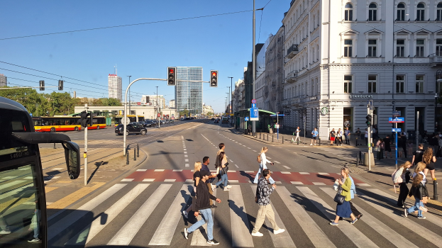 Eine Gruppe von Menschen, die eine Straße an einer Ampel überqueren, mit einem Bus auf der linken Seite, Fahrzeugen auf der Straße, Verkehrsampeln, Laternenmasten, Gebäuden, Bäumen und einem klaren blauen Himmel.