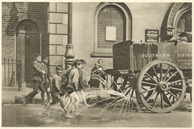 Schwarzes und wei├čes Bild von Kindern, die mit einem Hydranten vor einem Geb├Ąude mit Fenstern, einer Tür, einem Mast und einem Zaun spielen, wobei im Hintergrund ein Wagen zu sehen ist.