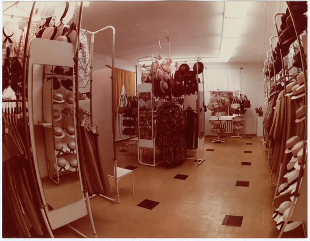 Altes Schwarz-Weiß-Foto eines kleinen Bekleidungsgeschäfts-Innenraums mit Kleiderstangen, Hutständern, einem Spiegel, einem Stuhl, einem Tisch mit diversen Gegenständen, Vorhängen und Deckenleuchten.