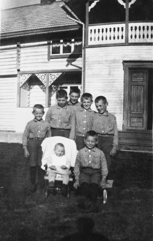 Ein Schwarz-Weiß-Foto einer Gruppe von Kindern, die vor einem Haus stehen, mit einem Baby im Kinderwagen in der Mitte.