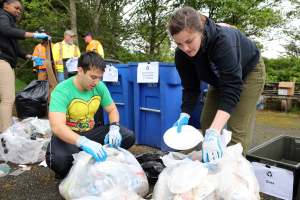 Zwei Personen in Handschuhen sammeln Müll auf Tellern in einem Park, umgeben von weggeworfenem Material, mit einem Mülleimer und einer Bank in der Nähe unter Bäumen und einem klaren Himmel.