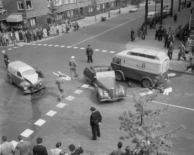 Schwarz-weiß-Szene eines Autounfalls an einer Straßenkreuzung mit einem Van, anderen Fahrzeugen und einer Gruppe von Menschen, mit Gebäuden, Bäumen, Pfählen und Texttafeln im Hintergrund.