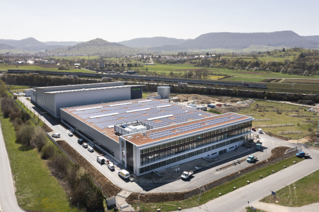 Eine Luftaufnahme eines großen Gebäudes mit Solarmodulen auf dem Dach, umgeben von Fahrzeugen, Bäumen und Masten, mit Hügeln und einem klaren blauen Himmel im Hintergrund, das die erste Fabrik in Deutschland zeigt, die mit Solarenergie gebaut wurde.