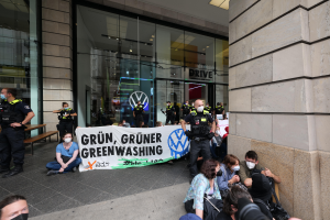 Eine Gruppe von Menschen sitzt vor einer Volkswagen-Filiale auf dem Boden und hält ein Transparent mit Text, einige tragen Masken und andere halten Kameras, mit Bänken und einem Gebäude mit Glaswänden im Hintergrund und Lampen an der Decke.