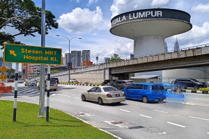 Eine belebte Straße mit Autos, eine Brücke im Hintergrund, Straßenlaternen, ein Schild, Verkehrskegel, Gras, Bäume, Gebäude und ein bewölkter Himmel.