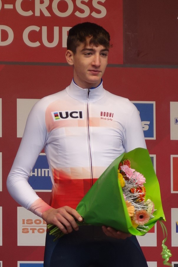 Ein Mann in einem Anzug steht auf einem Podium und hält einen Blumenstrauß, feiernd mit einem selbstbewussten und stolzen Gesichtsausdruck.