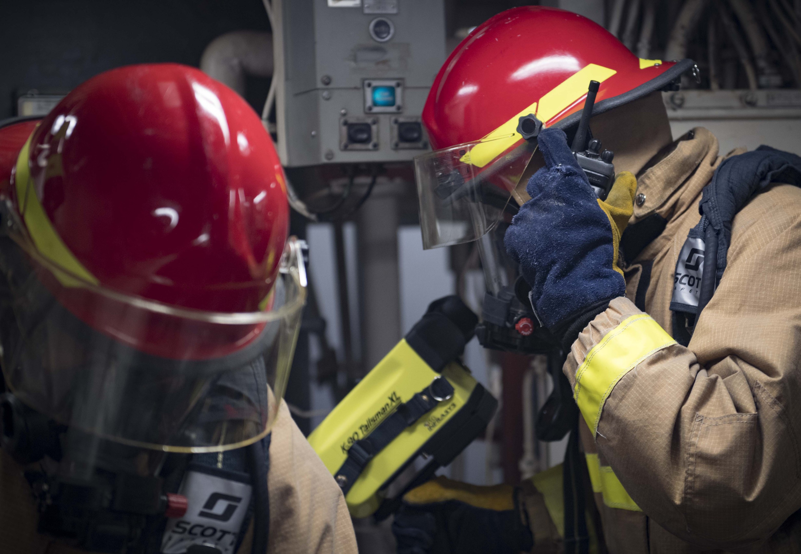 Zwei Feuerwehrleute in Schutzausrüstung arbeiten an einem Feuerhydranten während einer Übung mit Maschinen und Kabeln im Hintergrund.