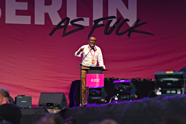 Mann in weißem Hemd und braunen Hosen steht auf einer Bühne und hält ein Mikrofon, mit einem pinken Banner im Hintergrund, das 'Berlin as Fuck' trägt, und Lautsprechern und anderen Gegenständen auf der Bühne.