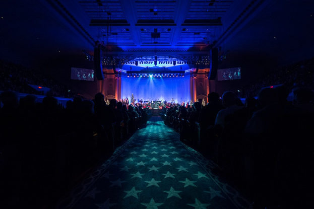 Ein großer Auditorium mit sitzenden Zuschauern, eine Bühne mit Musikern und über Kopfbeleuchtung und Bildschirme, was auf einen Konzertsaal während der Nationalen Convention 2019 hindeutet.