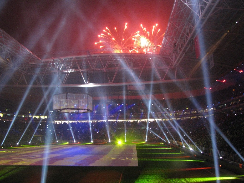 Das Olympische Stadion in London bei Nacht, erleuchtet mit hellen Lichtern und Feuerwerk, mit einem großen Bildschirm im Vordergrund und einer Menge drinnen.