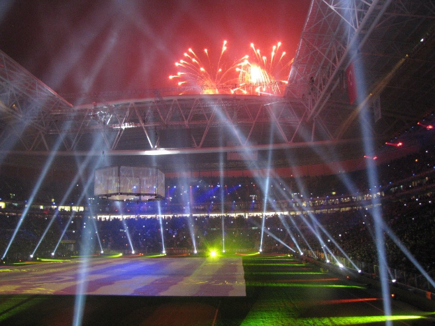 Das Olympische Stadion in London bei Nacht, erleuchtet mit hellen Lichtern und Feuerwerk, mit einem großen Bildschirm im Vordergrund und einer Menge drinnen.