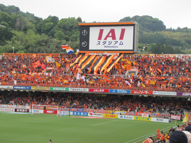 Fussballspiel in einem Stadion mit einer grossen Zuschauermenge, saftigem Grün, Tor, Bannern, Fahnen, einem grossen Bildschirm, Bäumen und einem klaren blauen Himmel.