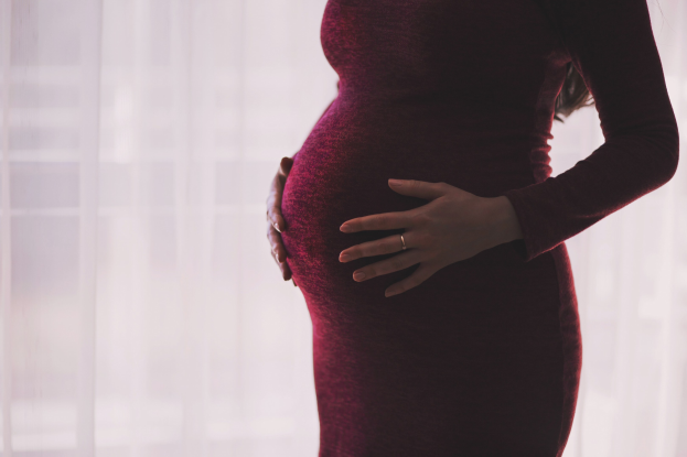 Eine schwangere Frau in einem roten Kleid steht neben einem Fenster, Hände auf dem Bauch, mit einem leicht unscharfen Hintergrund.