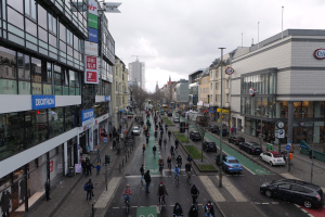 Eine belebte Stadtstraße mit Fußgängern, Radfahrern und Fahrzeugen, gesäumt von glaswandigen Gebäuden, Laternen und Schildern, unter einem klaren blauen Himmel mit Bäumen im Hintergrund.