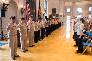 Eine Gruppe uniformierter Personen steht in einem Raum mit Stühlen auf der rechten Seite, Fotorahmen an der Wand, Kerzen und einem Lautsprecher auf der linken Seite und einer Fahne in der Mitte, alle salutierend während einer Militärzeremonie.