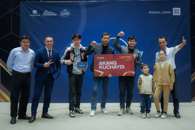 Gruppe von Menschen auf einer Bühne, die eine Tafel mit dem Text 'Akang Kuchaydi - Zakovat Quiz' halten, mit einem Banner, einem Barcode und einer Hintergrundbeleuchtung.