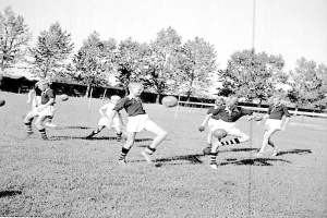 Schwarzes und weißes Bild von jungen Jungs, die auf einem Feld mit Bäumen und einem Pfosten im Hintergrund Fußball spielen.