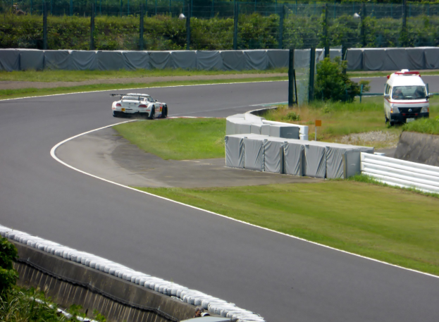 Ein Rennauto fährt auf einer Rennstrecke mit einem Polizeiwagen im Hintergrund, umgeben von Gras, Pflanzen, Bäumen und einem Zaun, mit einem Fahrzeug auf der rechten Seite und Text unten.