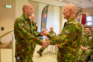Zwei Männer in Militäruniformen geben sich die Hand vor einer sitzenden Gruppe, mit einem Tisch, Geländer, Fahnen und diversen Hintergrundelementen einschließlich einer Tür, einer Schautafel, einem Fenster mit Vorhang und Deckenleuchten.