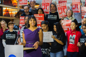 Eine Black-Lives-Matter-Aktivistin spricht in ein Mikrofon auf einem Podium, während sie eine Menge anspricht, die Schilder hält, mit Scheinwerfern und Gegenständen im Hintergrund.