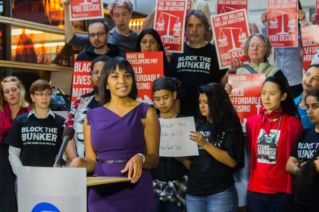 Eine Black-Lives-Matter-Aktivistin spricht in ein Mikrofon auf einem Podium, während sie eine Menge anspricht, die Schilder hält, mit Scheinwerfern und Gegenständen im Hintergrund.