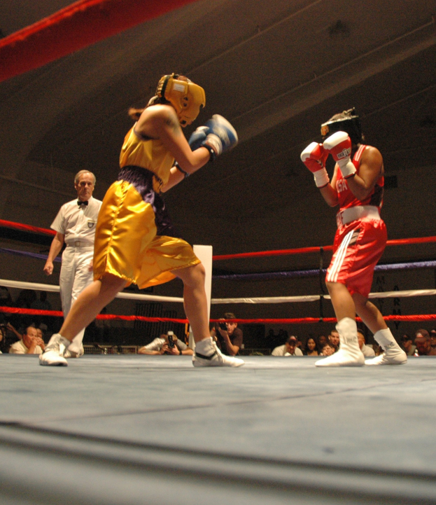 Zwei Boxer kämpfen im Ring mit einem Schiedsrichter und Zuschauern, einige Fotografen sind anwesend, Decke oben sichtbar.