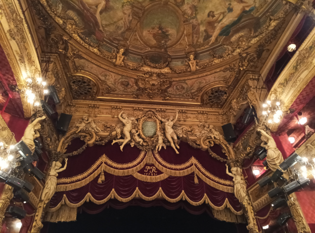 Innensicht eines Theaters mit roter Gardine, Kronleuchtern, Skulpturen, Lichtern, einer Deckenmalerei und einer Bühne mit einigen Menschen unten.