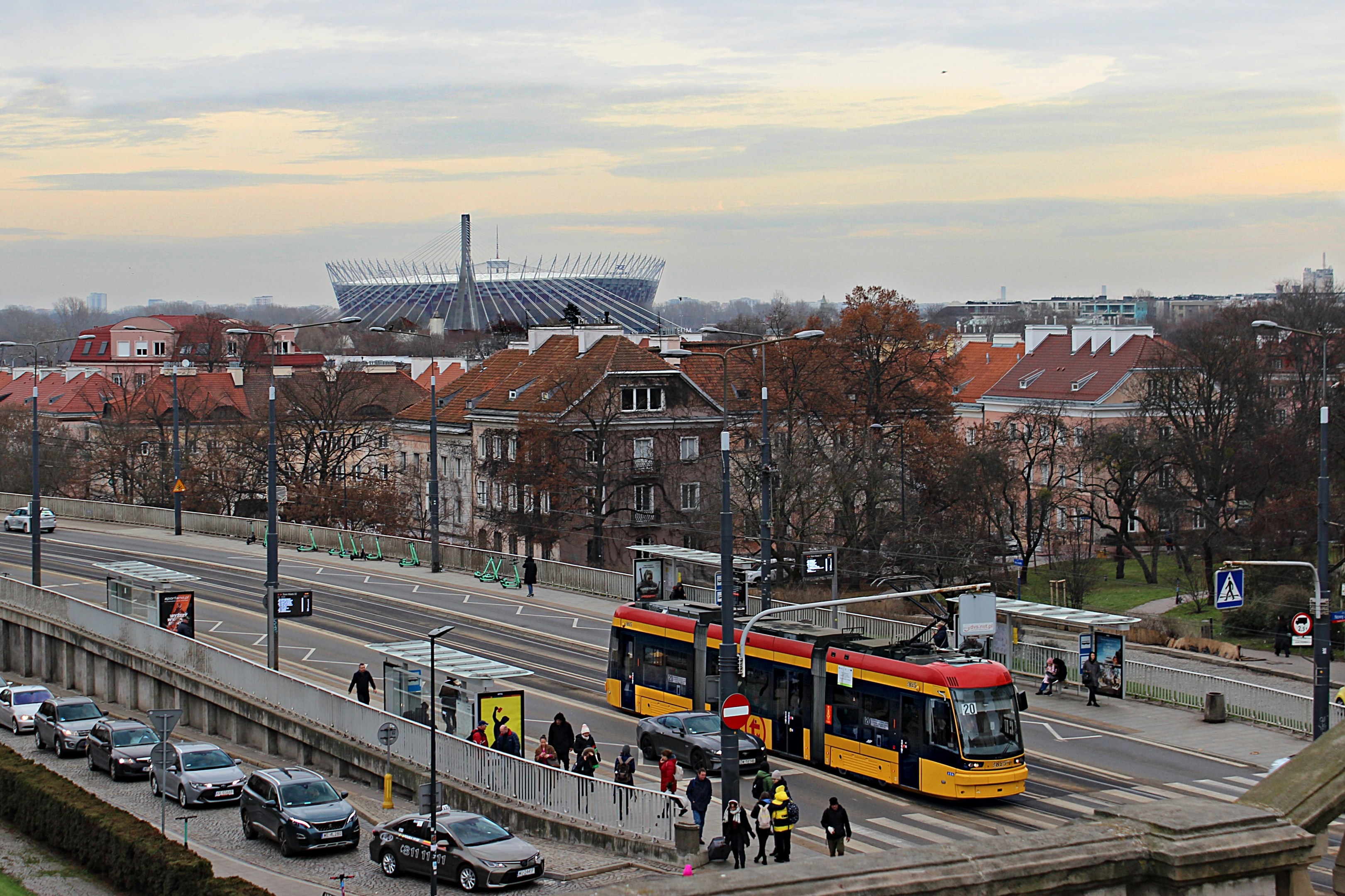 Eine gelbe und schwarze Straßenbahn fährt eine belebte Stadtstraße entlang, die von hohen Gebäuden, Autos, Fußgängern, Laternen, Schildern, Bäumen und Gras gesäumt ist, mit bewölktem Himmel im Hintergrund.