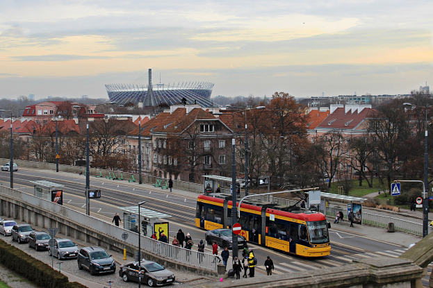 Eine gelbe und schwarze Straßenbahn fährt eine belebte Stadtstraße entlang, die von hohen Gebäuden, Autos, Fußgängern, Laternen, Schildern, Bäumen und Gras gesäumt ist, mit bewölktem Himmel im Hintergrund.