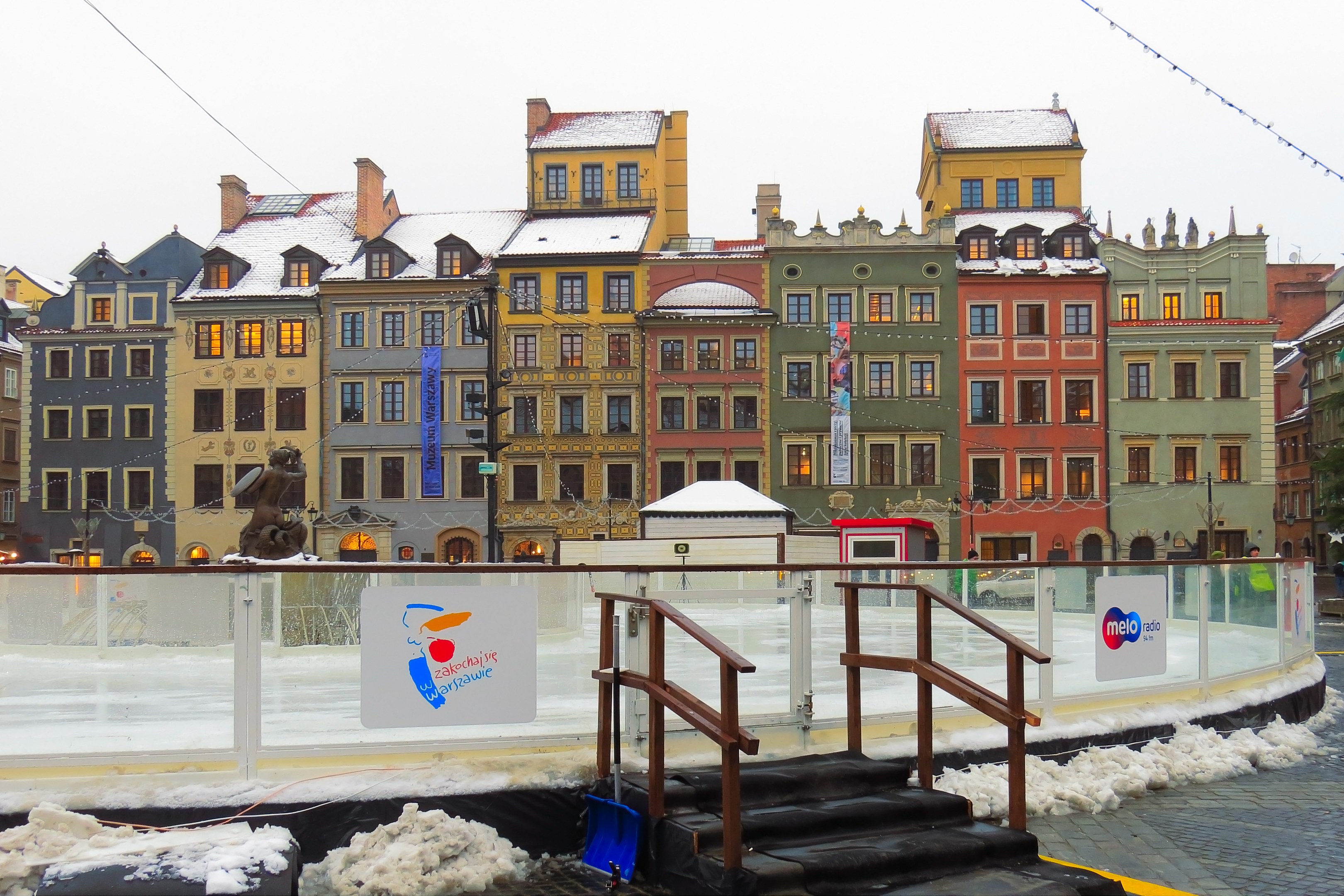 Eisbahn in einem Stadtplatz umgeben von Gebäuden, Treppen, Geländern, Informationsschildern, schneebedecktem Boden, Laternen, Bäumen und einem Himmelhintergrund.
