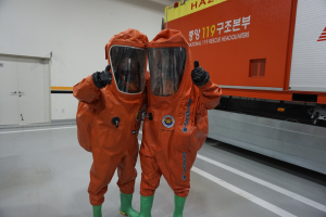 Zwei Personen in orangen Schutzanzügen stehen nebeneinander vor einem LKW, mit einer Wand und Türen im Hintergrund, was darauf hinweist, dass sie Teil des Nationalen Einsatzteams sind.