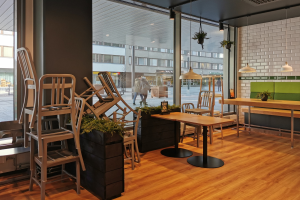 Ein Restaurantinnenraum mit hölzernen Tischen und Stühlen, großen Fenstern und Indoor-Pflanzen, mit Blick auf eine Berliner Straße mit Fußgängern, Gebäuden und Straßenelementen.