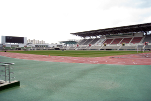 Ein großes Stadion mit einer Laufbahn, einem Geländer auf der linken Seite und Gebäuden unter einem klaren blauen Himmel im Hintergrund.