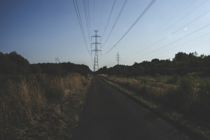 Eine Straße mit Stromleitungen, die entlang verläuft, umgeben von saftigem Grün und Bäumen, mit hohen Türmen und Drähten im Hintergrund unter einem klaren blauen Himmel.