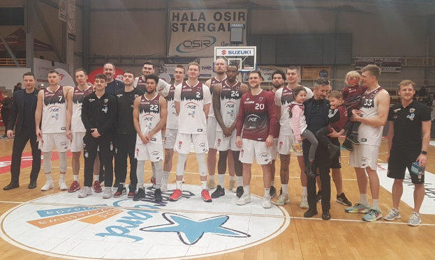 Gruppe von Männern und Kindern auf einem Basketballfeld mit Schildern "Hala Osir Starga", sitzende Zuschauer, ein Basketballkorb, Metallstangen, Deckenbeleuchtung und eine Wand.
