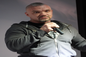 Ein Mann in einer schwarzen Kapuzenjacke und einem schwarzen Anzug mit weißem Hemd hält ein Mikrofon und steht selbstbewusst mit zurückgekämmtem Haar vor einer Wand.