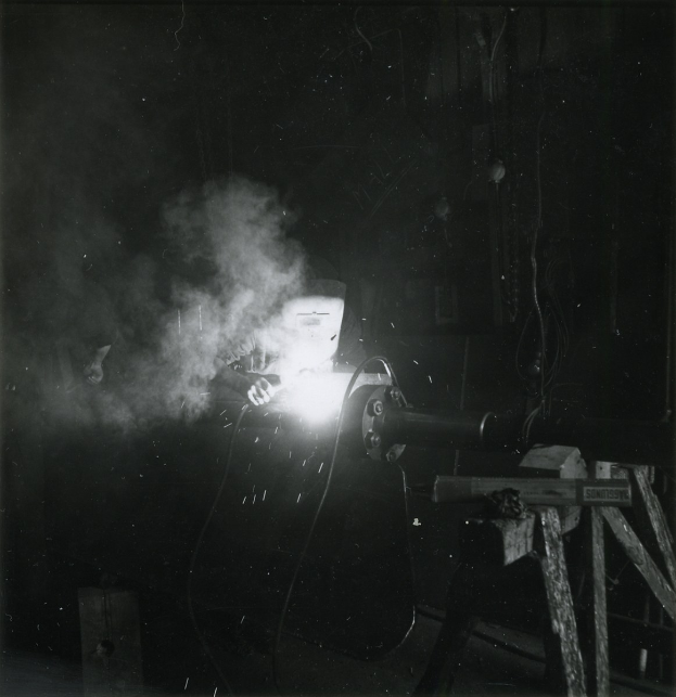 Schwarzes und weißes Foto eines Schweißers bei der Arbeit an Metall, mit Rauch aus der Schweißmaschine und sichtbaren Hintergrundobjekten.