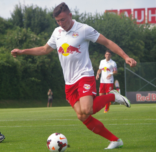Ein professioneller Fußballspieler in einem roten Trikot schießt einen Ball auf einem grünen Feld mit Bäumen und einem klaren blauen Himmel im Hintergrund, mit "RB Leipzig" auf einer Tafel.