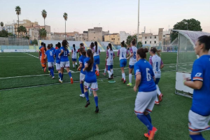 Eine Gruppe von Frauen, die auf einem Outdoor-Feld Fußball spielen, mit einem Torpfosten auf der rechten Seite, umgeben von Gebäuden, Bäumen und einem klaren blauen Himmel.