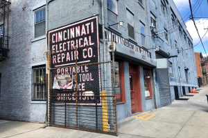 Das Cincinnati Electrical Repair Co. Gebäude mit Fenstern, Türen, einem Schild, einem Metallzaun, einem Müllcontainer, Polen, Drähten, Bäumen und einem bewölkten Himmel.