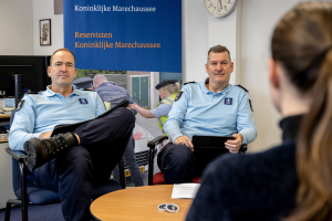 Zwei Polizeibeamte sitzen an einem Tisch mit einer Frau, umgeben von Dokumenten und Gegenständen, mit einem Wandbanner, einer Uhr und Fotorahmen im Hintergrund und einem Computersystem auf einem separaten Tisch.