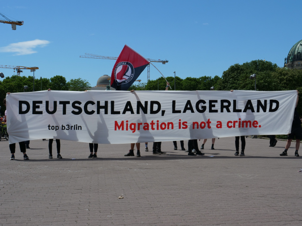 Menschen mit einem Transparent mit der Aufschrift "Deutschland, Lagerland Migration ist kein Verbrechen" und einer Flagge, im Hintergrund Straßenlaternen, Straßenmasten, Bäume, Kräne, ein Gebäude und ein bewölktes Himmel.